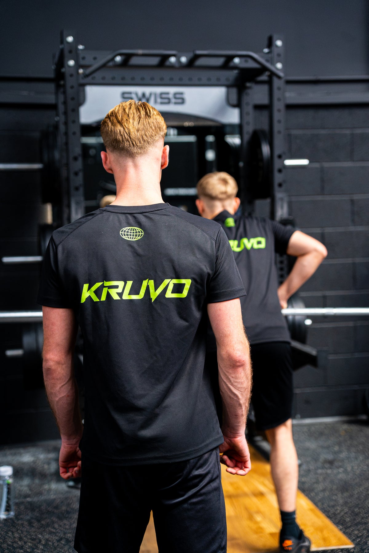 Athlete seen from behind in a black KRUVO T-shirt at a squat rack, with a training partner beside him