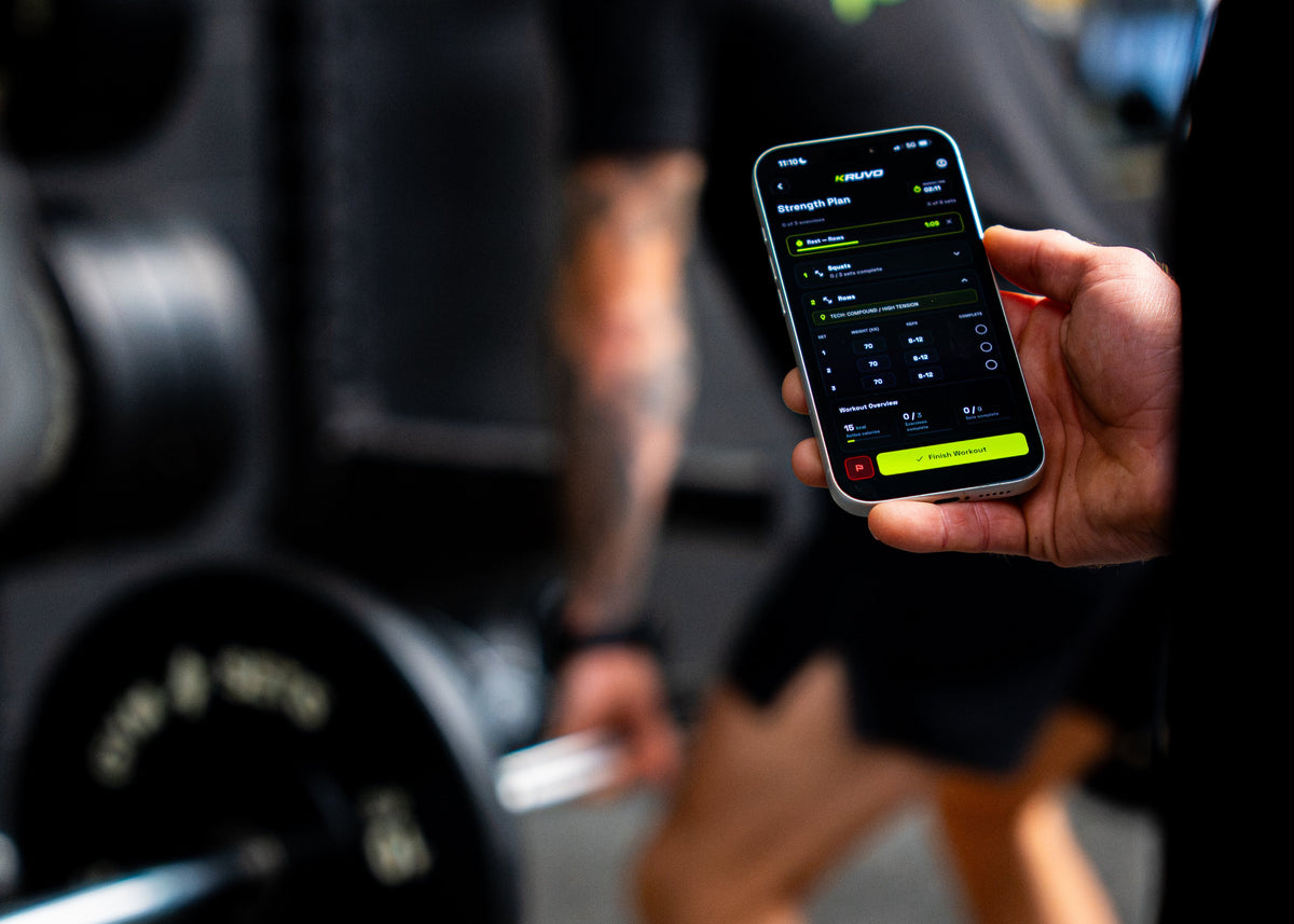 Hand holding a phone showing a training plan in the KRUVO app, with a barbell in the background