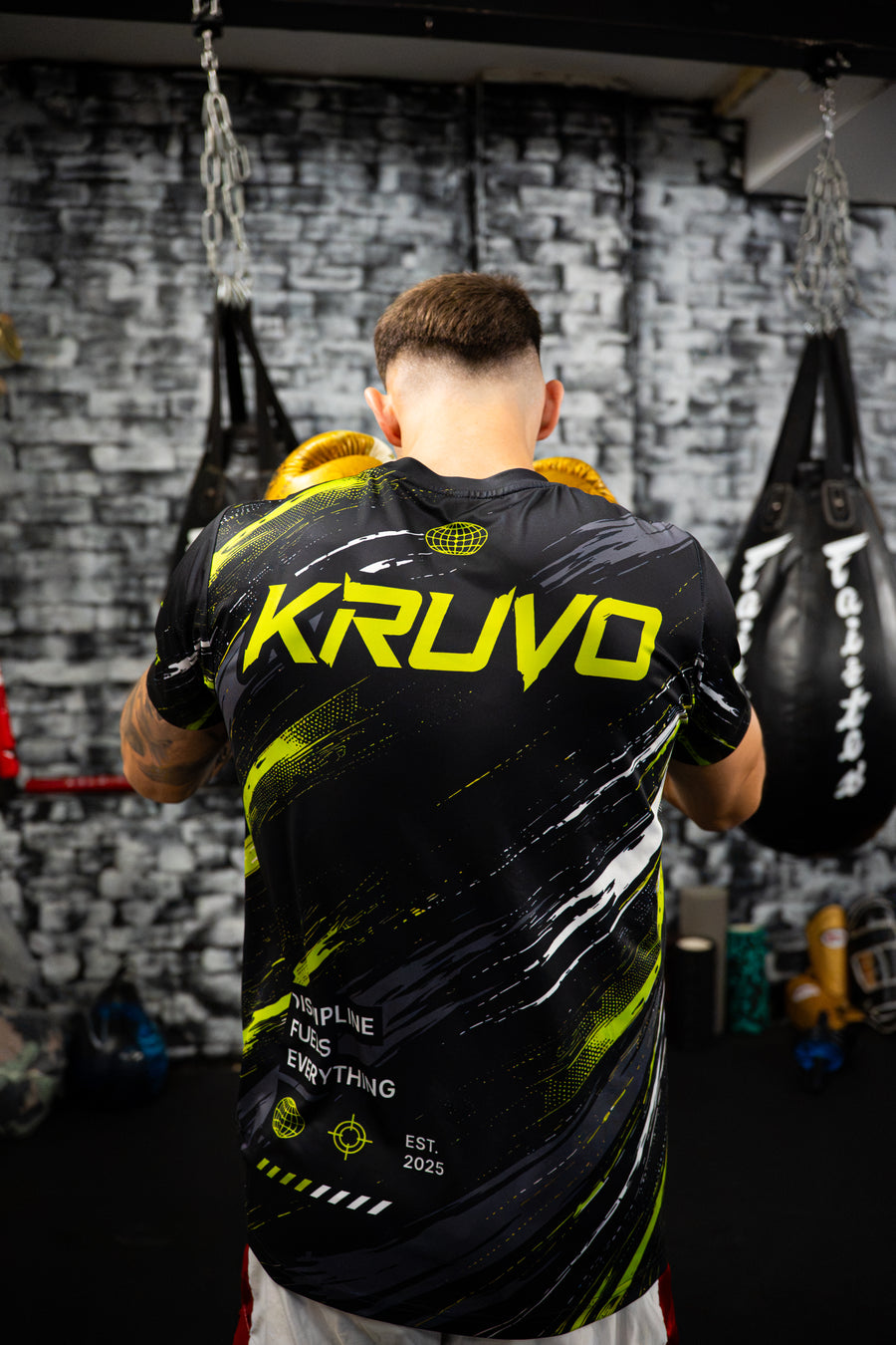 Fighter seen from behind in a black and neon green KRUVO jersey, standing between heavy bags in the gym